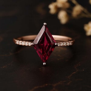 Peut inclure: Une bague en or rose ornée d'une pierre précieuse rouge foncé en forme de losange. La monture est ornée de petits diamants transparents. La bague est posée sur un fond sombre et texturé, avec des accents floraux flous.