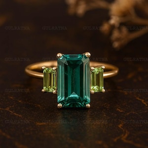May include: A gold ring with a central emerald-cut teal gemstone and two smaller, rectangular green gemstones. The ring has a slender band. The image shows the word "GULRATNA". The ring is set against a dark background.