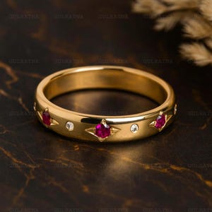 May include: A gold band ring with a series of alternating ruby and diamond accents. The rubies are a deep red color and are set in a star-shaped design. The diamonds are small and round. The ring is on a dark background.