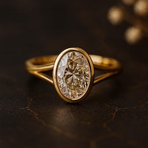 May include: A gold ring with an oval-shaped diamond. The diamond is set within a bezel, and the band has a split shank design. The ring is shown against a dark backdrop, accentuating the gemstone's brilliance.