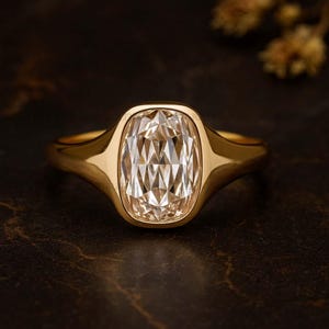 Puede incluir: Un anillo de oro con una gran gema clara de corte rectangular. La gema está engastada en un bisel de oro liso. La banda del anillo también es de oro y tiene un diseño redondeado. El anillo se muestra sobre una superficie oscura y texturizada.