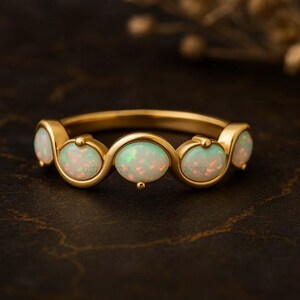 May include: A gold ring featuring five oval opal gemstones. The opals have a milky white base with flashes of color. The ring's band is a smooth, polished gold, and the opals are set in a delicate, curved design.