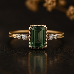May include: A gold ring with a rectangular, emerald-cut green gemstone. The ring is embellished with small, round diamonds on either side of the main stone. The band has a delicate, beaded design.