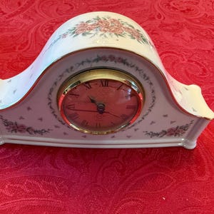 Puede incluir: Un reloj de chimenea de porcelana blanca con esfera dorada y números romanos. El reloj está decorado con motivos florales rosas y ribetes dorados. El reloj mide aproximadamente 20 cm de ancho.