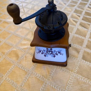 May include: A vintage coffee grinder with a wooden base and handle, and a black metal grinding mechanism. The base features a white ceramic drawer with a floral design in shades of purple and blue.