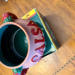 May include: A ceramic salsa bowl with a light pink exterior and a green interior. The word "SALSA" is written in red around the bowl. The bowl has two red chili pepper handles and sits next to a green and yellow box.