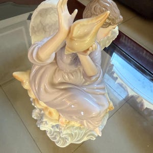 May include: A porcelain figurine of an angel, holding a conch shell. The angel is dressed in a lavender gown and has feathered wings. The figurine is resting on a cloud-like base, with a soft, pastel color palette.