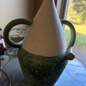 Puede incluir: Un jarrón de cerámica con una sección superior blanca y una sección inferior verde moteada. El jarrón tiene dos asas verdes y un pequeño acento verde redondo. El jarrón está sobre una mesa de cristal.