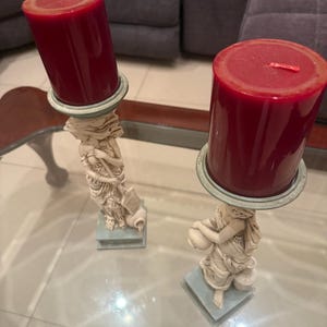 May include: Two decorative candle holders with burgundy pillar candles. The candle holders feature a classical female figure design in off-white and pale blue. The candles are cylindrical and have a single wick.