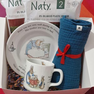 May include: A gift set in a white box featuring a plate and mug with Peter Rabbit illustrations, a blue washcloth tied with a red ribbon, and two packs of baby diapers. The plate has text around the rim.
