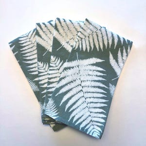 May include: Four folded cloth napkins with a fern leaf pattern. The napkins are a muted teal color with white fern leaf designs. The napkins are stacked on a white surface.