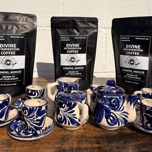 Könnte beinhalten: Drei schwarze Beutel "Divine Intervention Coffee" aus Chiapas, Mexiko, mit einem passenden blau-weißen Keramik-Kaffeeservice. Das Set umfasst Tassen, Untertassen, einen Sahnekännchen und eine Teekanne, alle mit detailliertem Blumendesign.