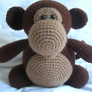 Pode incluir: Um brinquedo de macaco de crochê marrom com olhos pretos. O macaco está sentado em uma superfície branca.
