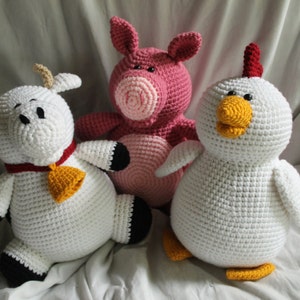 Peut inclure: Trois animaux en peluche crochetés : une vache blanche avec un collier rouge et jaune, un cochon rose et un poulet blanc avec une crête rouge.