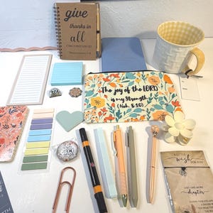 May include: A collection of stationery and accessories, including a weekly planner, notebooks, pens, and a floral-patterned pouch with the text "The joy of the LORD is my Strength." A mug and a decorative flower are also present.