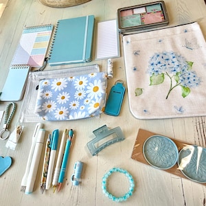 May include: An assortment of stationery and accessories in shades of blue and white. Items include notebooks, pens, a daisy-print pouch, a floral-print bag, a hair clip, coasters, and a beaded bracelet. The items are arranged on a light-colored wooden surface.