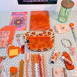May include: A collection of orange stationery and accessories, including notebooks, pens, a pencil, a scrunchie, a water bottle, and a coloring book. The items are arranged on a patterned surface, creating a visually appealing collection.