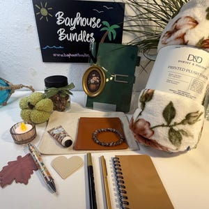 May include: A collection of items including a D&D printed plush throw, a green journal with an oval floral pendant, a small jar with a green ribbon, a tan notebook, two pens, a beaded bracelet, a heart-shaped tag, a leaf cutout, and a candle.