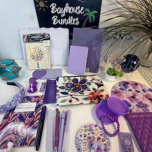 May include: A collection of purple-themed items including a Bayhouse Bundles sign, note cards with a tree design, journals, a floral pouch, pens, a headband, a beaded bracelet, a hand cream tube, a quilted case, and various decorative accessories.