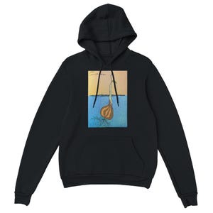 Op de afbeelding: Zwarte hoodie met een grafische print van een ui met wortels, een stengel en een blad. De afbeelding staat tegen een blauwe en gele achtergrond. De tekst in de afbeelding is wazig.