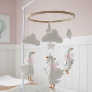 Pode incluir: Móvel de berço com gansos de feltro branco com laços rosa, nuvens, estrelas e luas, suspensos por um anel de madeira. Pendurado sobre um berço branco num quarto rosa.
