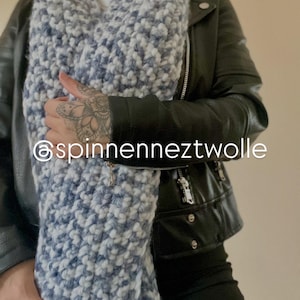 May include: A chunky knit scarf in shades of blue and white. The scarf is thick and textured, with a fringed edge. The text "@spinnenneztwolle" is visible on the scarf. The scarf is worn over a black leather jacket.