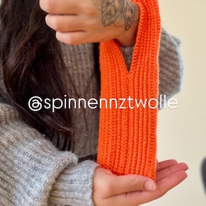Puede incluir: Manga de punto naranja brillante. La manga está hecha de un hilo grueso y texturizado. El texto "@spinnennztwolle" es visible. Un accesorio llamativo y vibrante.