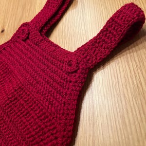 Puede incluir: Primer plano de un artículo rojo tejido a mano con dos correas y detalles de botones. El artículo está hecho de hilo grueso y texturizado, posiblemente una prenda o parte de ella. El fondo es una superficie de madera clara.