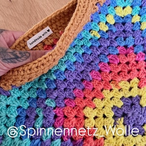 May include: A handmade, colorful crochet item with a granny square design. The item features a vibrant mix of rainbow colors, including blue, purple, pink, yellow, and orange. The border is a warm brown color. The text "handmade with" is visible on a small tag.