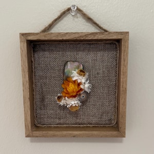 May include: Square wooden framed art piece with a burlap backing. The artwork features a cluster of dried flowers in shades of orange, white, and yellow. A natural twine loop is attached for hanging. The frame is approximately 6 inches square.