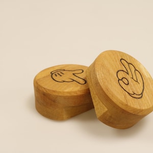 Könnte beinhalten: Zwei runde Holzboxen mit Handgesten-Designs. Eine Box zeigt eine Hand, die ein Friedenszeichen macht, die andere eine Hand, die eine „Okay“-Geste macht. Die Boxen sind aus hellem Holz gefertigt.