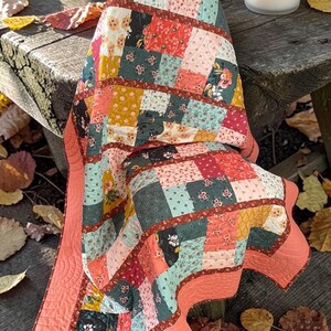 May include: A patchwork quilt draped over a wooden bench. The quilt features a variety of floral and geometric patterns in shades of coral, green, yellow, and red. The quilt's border is a solid coral color. A lit candle sits nearby.