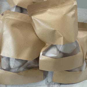 May include: Several brown paper pouches with clear window panels are stacked together. The pouches have a zip-top closure and are filled with a white substance. The pouches are sitting on a white surface.