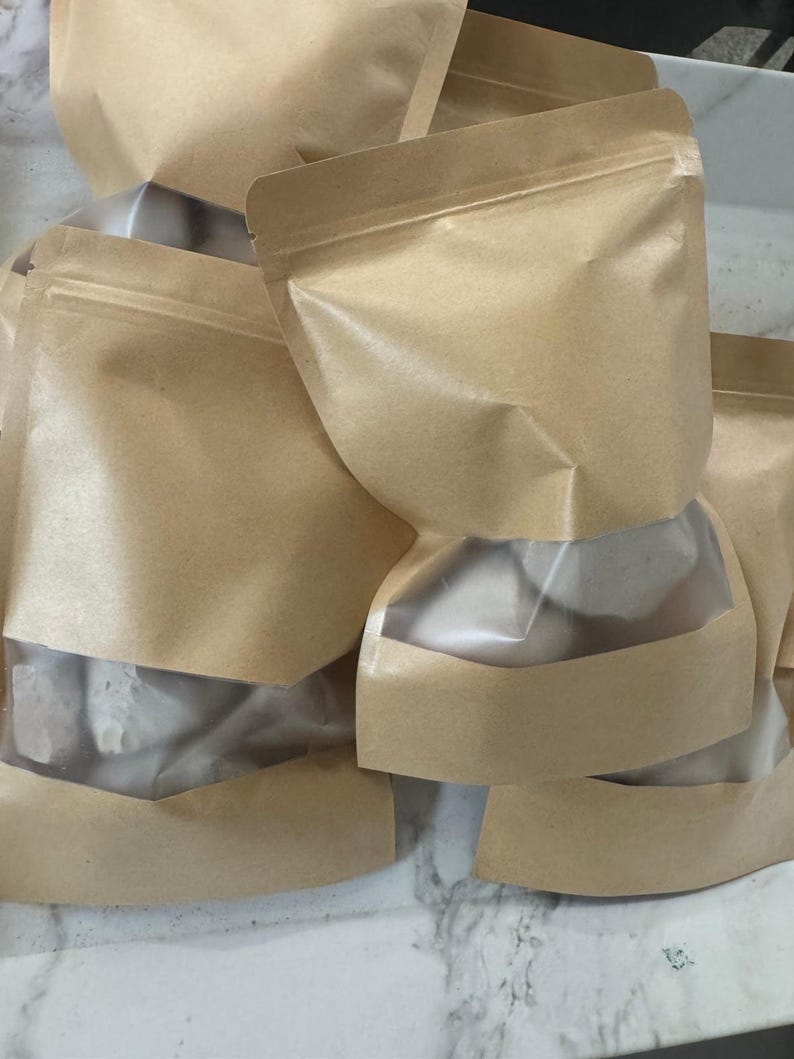 May include: Several brown paper pouches with clear window panels are stacked together. The pouches have a zip-top closure and are filled with a white substance. The pouches are sitting on a white surface.