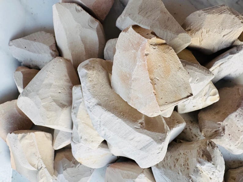 May include: A pile of light beige, irregularly shaped chalk pieces. The chalk appears to be roughly cut, with visible texture and variations in color. The pieces are stacked together, creating a natural, earthy aesthetic.