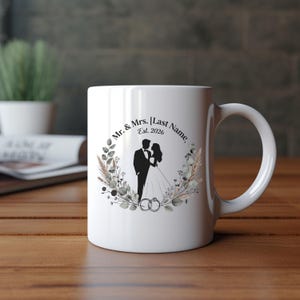 Könnte beinhalten: Eine weiße Keramiktasse mit einer schwarzen Silhouette eines Paares in Hochzeitskleidung, umgeben von einem botanischen Kranz aus getrockneten Blättern und Blumen. Der Text lautet Mr. & Mrs. [Last Name] Est. 2026, mit zwei ineinander verschlungenen Ringen.