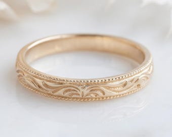 May include: A gold ring featuring a delicate engraved scroll pattern along the outer band, bordered by fine milgrain detailing on both the top and bottom edges. The metal has a warm, polished finish that reflects light across its surface.