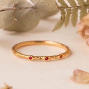 May include: A thin gold ring with a textured, organic band featuring three small, round, vibrant red gemstones set at intervals along the front. The metal surface has a slightly uneven, hammered finish, giving it a natural, twig-like appearance.