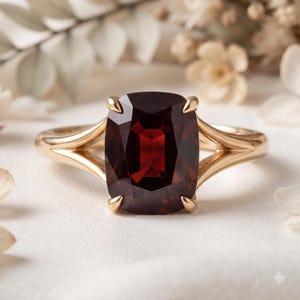 May include: A gold ring featuring a large, cushion-cut deep red gemstone held by four prongs. The band has a split-shank design that tapers toward the center stone, creating a sleek and polished look for this piece of fine jewelry.