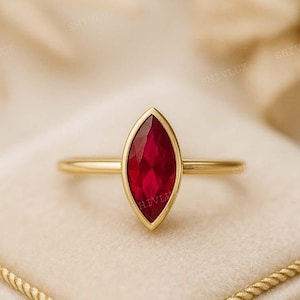 May include: A gold ring with a marquise-cut, deep red gemstone. The ring is set on a cream-colored, velvet-like surface, with a gold rope-like border. The ring's band is thin and simple.