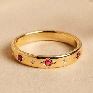 May include: A gold band ring featuring red gemstones in star settings, interspersed with small clear stones. The ring has a polished finish and is presented on a neutral background. The ring's design is symmetrical.
