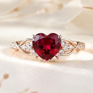 May include: A rose gold ring featuring a central heart-shaped deep red gemstone. The band is accented with small, clear, round-cut stones set along the sides, with an intricate, interwoven metal design leading up to the main heart-shaped stone.