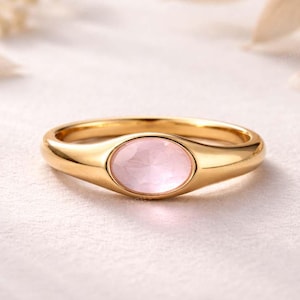 Op de afbeelding: Een gouden ring met een ovale, lichtroze edelsteen. De ring heeft een gladde, gepolijste band en een bezel-zetting voor de edelsteen. De achtergrond is een zachte, neutrale kleur, die het ontwerp benadrukt.