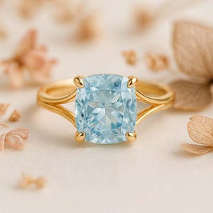 Puede incluir: Un anillo de oro con una gran gema de talla cojín de color azul claro. El anillo tiene un diseño de caña dividida y cuatro garras que aseguran la gema. El fondo es de un color suave y neutro con acentos florales borrosos.
