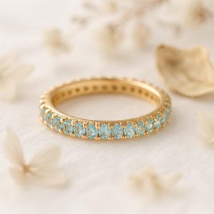 May include: A gold eternity ring featuring a continuous line of light blue gemstones. The ring is set against a soft, neutral background with blurred floral accents, emphasizing the ring's delicate design and colour.