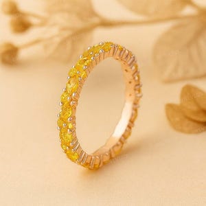 Op de afbeelding: Een goudkleurige ring met een band volledig bezet met ovaal geslepen gele edelstenen. De ring staat tegen een zachte, neutrale achtergrond, die het levendige geel van de stenen benadrukt. Het ontwerp van de ring is elegant en opvallend.
