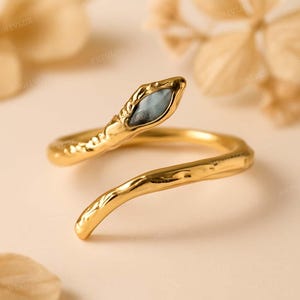 May include: A gold-colored snake ring with a textured finish. The snake's head features a blue-grey stone. The ring coils around the finger, creating a unique and stylish accessory. Jewelry.