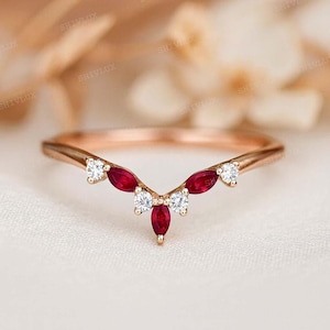 Könnte beinhalten: Roségoldener Ring mit geschwungenem Design. Der Ring hat kleine, runde, weiße Edelsteine und marquiseförmige, tiefrote Edelsteine. Der Ring ist vor einem weichen, neutralen Hintergrund platziert.