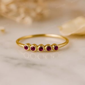May include: A delicate gold ring featuring five small, round, vibrant red gemstones. The ring has a thin band and the gemstones are evenly spaced across the top. The ring is set against a soft, blurred background.