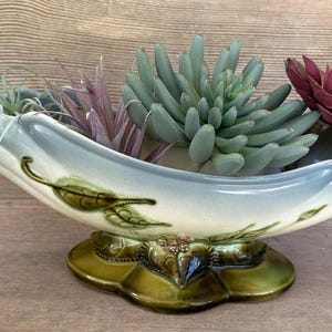 May include: A ceramic boat-shaped planter with a cream and light blue gradient finish, featuring embossed green leaf details and a dark green base. The planter is filled with a variety of artificial succulents in shades of green and deep red.